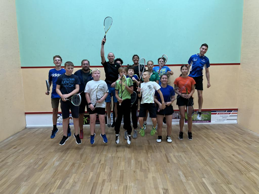 Kinder und Trainer springen in der Luft mit Schläger in der Hand auf einem Squash Court in der Sportscheune Weigmannsdorf